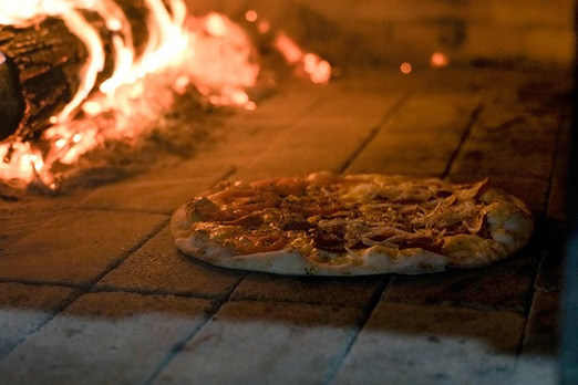 Pizza im Holzofen beim Backen mit offener Flamme und glühenden Kohlen im Hintergrund